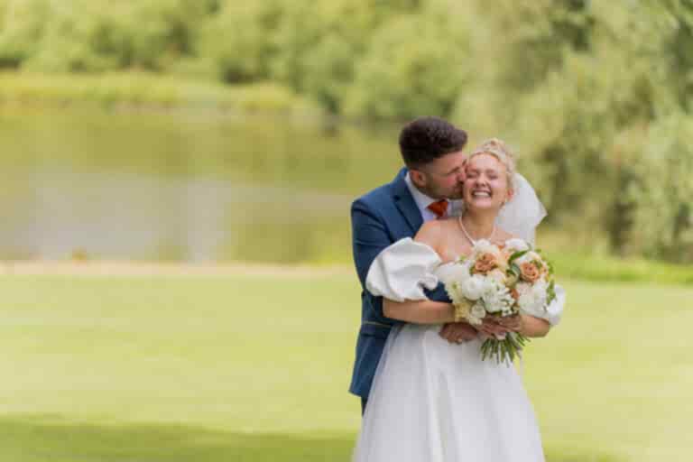 A bride and groom embrace in front of a lake, with the groom kissing the bride on the cheek, both smiling. The bride holds a bouquet of flowers and the background features greenery and water.
