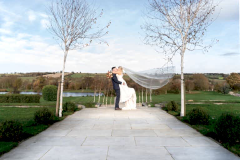 Bride and groom share a kiss outdoors, with the groom lifting the bride. Her veil flows behind as they stand on a stone path, surrounded by trees and a scenic landscape.